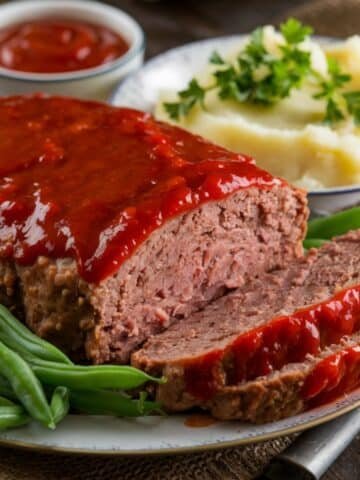 Juicy meatloaf recipe topped with a rich tomato glaze, served with creamy mashed potatoes, fresh green beans, and a bowl of ketchup on a rustic dining table.
