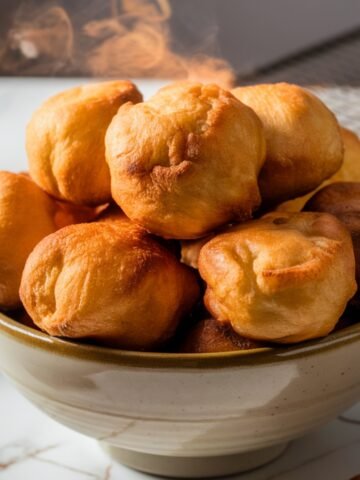 A bowl filled with freshly fried puff puff, a popular African deep-fried dough snack, with a golden-brown, crispy exterior. Steam rises from the warm puff puff, highlighting their soft and fluffy texture inside. The background features a cooling rack and a marble countertop, adding to the inviting presentation of this delicious Puff Puff Recipe.