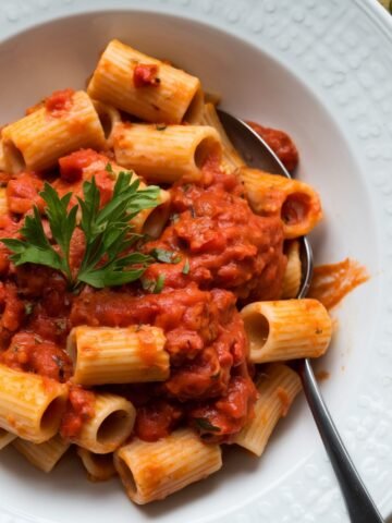A delicious plate of Ditalini Pasta Recipe (or rigatoni) coated in a rich, homemade tomato sauce, garnished with fresh parsley. Served in a white dish with a spoon, accompanied by crusty garlic bread. A classic Italian comfort food recipe.