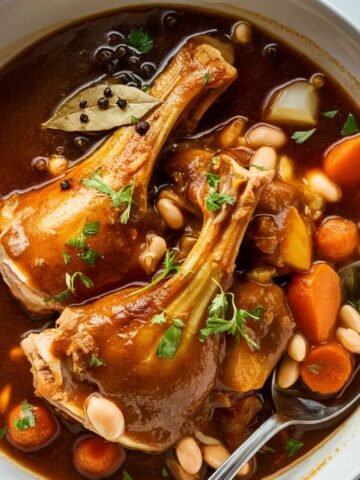 A hearty Turkey Neck Recipe featuring tender, slow-braised turkey necks in a rich, flavorful broth with carrots, onions, and fresh parsley. The dish is served in a rustic cast-iron skillet, showcasing golden-browned turkey necks partially submerged in a savory, aromatic sauce. The vibrant vegetables add color and depth, while the fresh herb garnish enhances the presentation. Warm natural lighting highlights the textures and flavors, making this dish a perfect comfort meal.