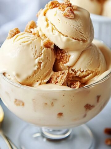A close-up of creamy homemade dulce de leche ice cream in a glass dish, topped with golden crumbles. The smooth scoops slightly drip down the sides, and a golden spoon is visible in the background.