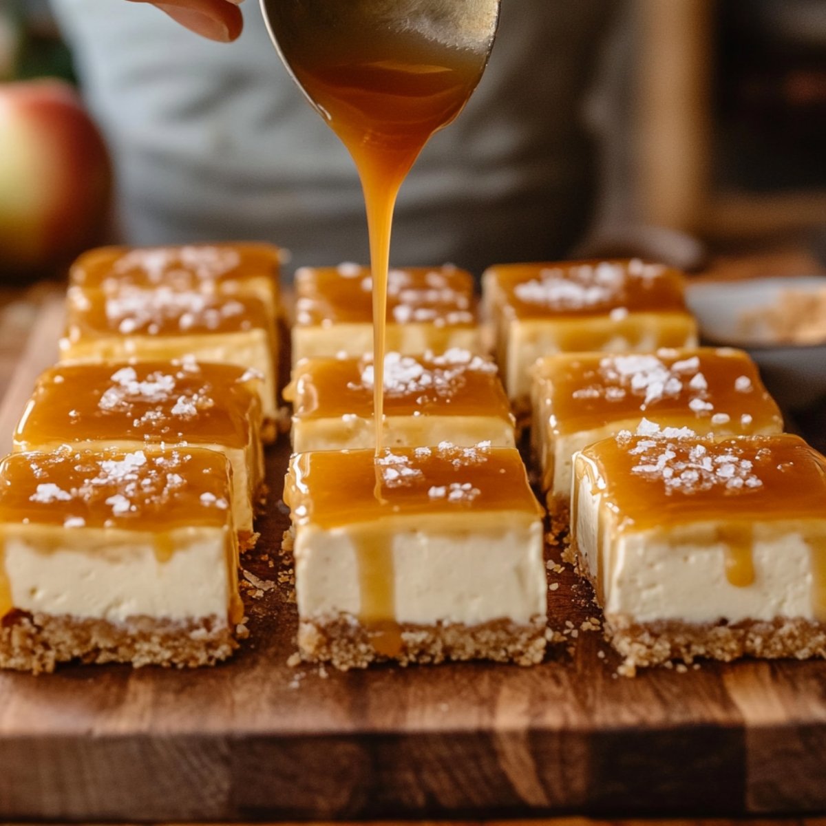 Homemade caramel apple cheesecake bars cut into squares on a wooden board, drizzled with caramel sauce and sprinkled with flaky sea salt, in warm natural lighting for a real, cozy feel.