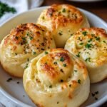 Homemade garlic cheese rolls arranged neatly in a white ceramic dish. The rolls appear golden-brown and freshly baked, topped generously with melted cheese, minced garlic, and finely chopped parsley. Each roll has a soft, fluffy texture with lightly crisp edges, displaying slight browning and bubbling cheese.