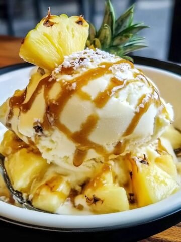 A close-up of homemade pineapple ice cream topped with caramel drizzle and fresh pineapple slices, resting on a bed of diced pineapple chunks. Soft natural light highlights the creamy texture and shiny caramel, with a spoon on the side.