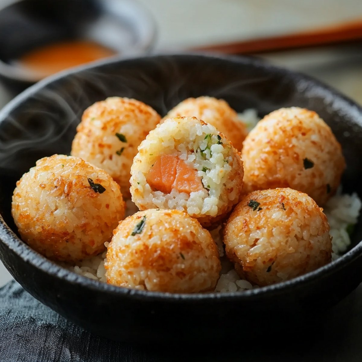 A bowl of homemade grilled salmon rice balls with a crispy golden crust, filled with tender cooked salmon, steaming hot and served in a rustic black bowl with dipping sauce and chopsticks in the background.