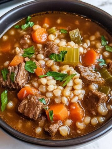 Homemade beef barley soup with tender beef, carrots, celery, and pearl barley in a rich broth, topped with fresh parsley in a black bowl.