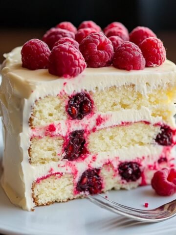 Homemade white chocolate raspberry cake slice with three moist layers, fresh raspberries, and creamy white chocolate frosting, served on a white plate with a fork.