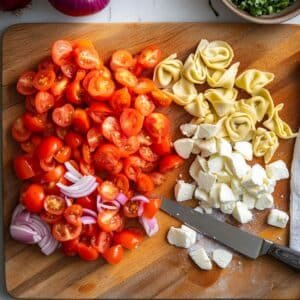 Chopped cherry tomatoes, red onion, mozzarella, and cheese tortellini on a wooden cutting board with a kitchen knife.