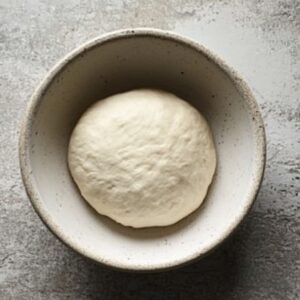 A ball of homemade pizza dough in a ceramic bowl on a gray countertop, ready to rise.