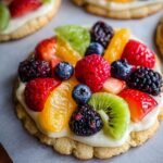 Homemade fruit pizza sugar cookie topped with creamy frosting and fresh berries, kiwi, and orange slices on parchment paper.