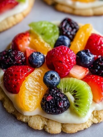 Homemade fruit pizza sugar cookie topped with creamy frosting and fresh berries, kiwi, and orange slices on parchment paper.