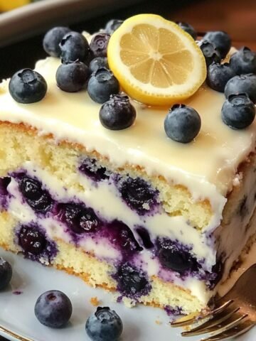 A homemade slice of lemon blueberry cake with white glaze, fresh blueberries, and a lemon slice on top, served on a white plate with a fork.