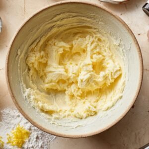 A mixing bowl with creamed butter and sugar, fluffy and pale yellow, with lemon zest and flour scattered on a marble countertop nearby.