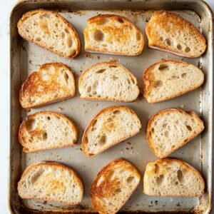 Toasted slices of rustic Italian bread on a worn baking sheet, golden and crisp with airy centers.