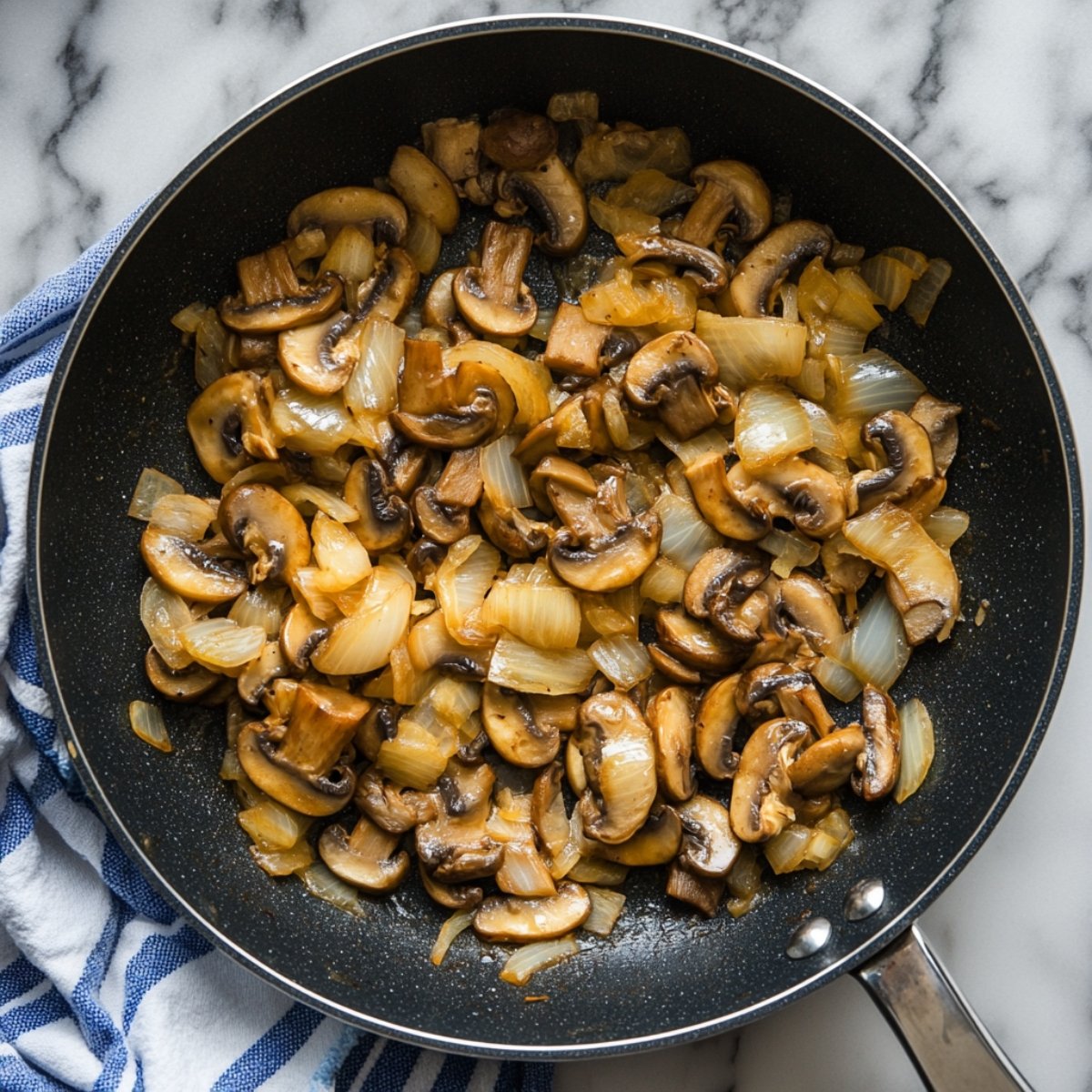 Sautéed mushrooms and onions in a black skillet, sitting on a marble counter with a blue-striped kitchen towel—part of the filling for beef Wellington.