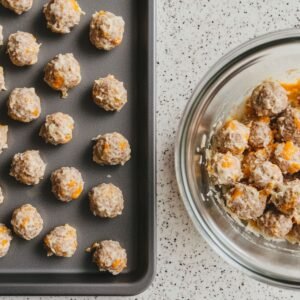 Unbaked sausage balls on a baking sheet beside a glass bowl with more rolled sausage balls, on a speckled countertop.