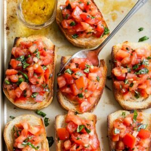 Toasted bread slices topped with fresh tomato, basil, and garlic mixture, with a spoon mid-assembly on a baking sheet.