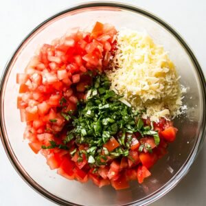Glass bowl with diced tomatoes, chopped basil, and shredded Parmesan, ready to mix for homemade bruschetta.