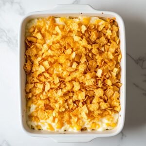 Unbaked hashbrown casserole in a white baking dish, topped with a layer of crushed golden cornflakes, sitting on a marble countertop.