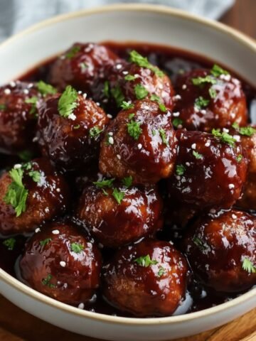 Homemade grape jelly meatballs coated in a glossy dark sauce, garnished with fresh parsley and a sprinkle of coarse salt, served in a white bowl.