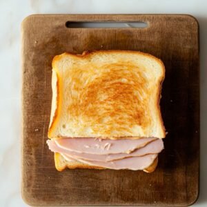 A toasted Monte Cristo sandwich with sliced ham, placed on a wooden cutting board, showing a golden, crisp top slice of bread with ham slightly sticking out.