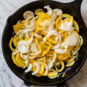 Sautéed yellow squash and sliced onions cooking in a cast iron skillet on a marble countertop, seasoned with salt and pepper.