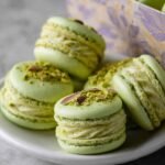 A plate of homemade pistachio macarons with light green shells and creamy piped filling, topped with crushed pistachios and pistachio slivers, with more macarons visible in a decorative gift box in the background.