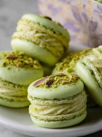 A plate of homemade pistachio macarons with light green shells and creamy piped filling, topped with crushed pistachios and pistachio slivers, with more macarons visible in a decorative gift box in the background.