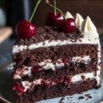 A slice of homemade Black Forest cake with chocolate layers, whipped cream, cherry filling, dark chocolate shavings, and fresh cherries on top.