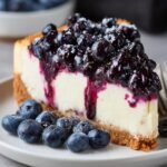Homemade blueberry cheesecake slice with a golden graham cracker crust, creamy filling, and glossy blueberry topping, served with fresh berries on a plate.