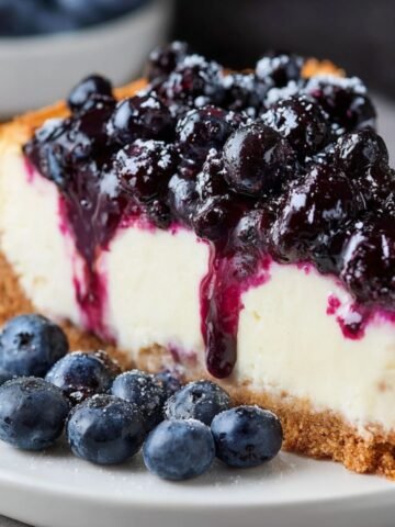 Homemade blueberry cheesecake slice with a golden graham cracker crust, creamy filling, and glossy blueberry topping, served with fresh berries on a plate.