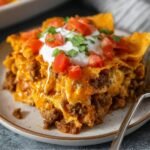 Homemade Dorito casserole slice topped with sour cream, diced tomatoes, and cilantro, served on a plate with melted cheese and crunchy chips visible.