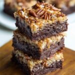 A stack of three thick, fudgy German chocolate brownies with gooey coconut-pecan topping, resting on a wooden board. The rich chocolate base looks moist and dense, with toasted pecans and shredded coconut scattered unevenly for a rustic, homemade touch.