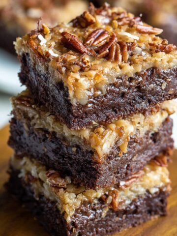 A stack of three thick, fudgy German chocolate brownies with gooey coconut-pecan topping, resting on a wooden board. The rich chocolate base looks moist and dense, with toasted pecans and shredded coconut scattered unevenly for a rustic, homemade touch.