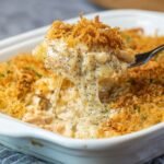 A fork lifts a creamy, cheesy scoop of homemade poppy seed chicken casserole from a white baking dish, showing tender chicken, black poppy seeds, and a golden, crispy topping.
