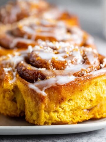Homemade pumpkin cinnamon rolls topped with cream cheese glaze, showing golden-orange dough swirls and gooey spiced filling on a white plate.