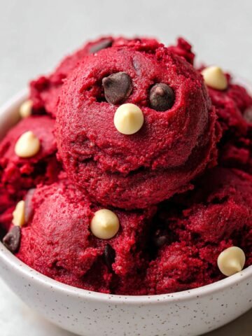 Homemade red velvet cookie dough scoops in a white bowl, studded with white and dark chocolate chips, on a light gray surface.