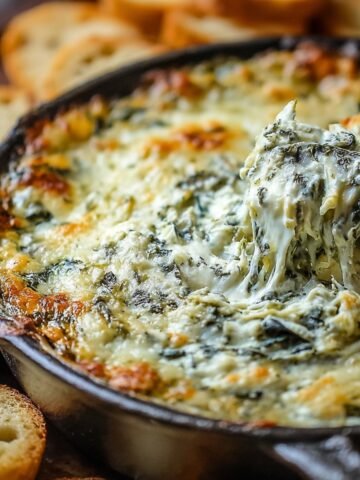 Homemade Spinach Artichoke Dip in a cast iron skillet, with a baguette slice lifting gooey, melty cheese, surrounded by toasted bread slices.