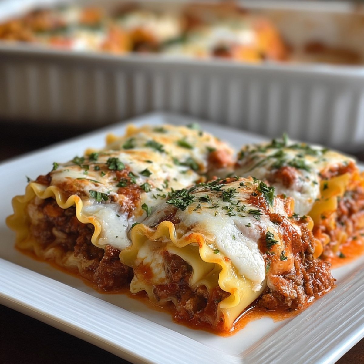 Four lasagna roll ups on a white plate, topped with melted cheese and parsley, with meat sauce visible. A baking dish appears blurred in the background.