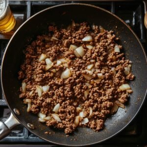 Top-down view of browned ground beef and sautéed onions in a skillet, cooked and ready for cheeseburger casserole. Warm, natural lighting gives it a homemade feel.