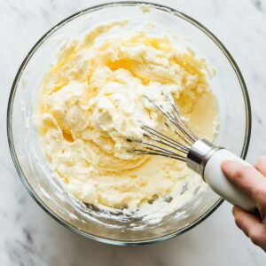 Glass bowl of creamed butter and cream cheese being whisked by hand on a marble countertop, part of red velvet cookie dough preparation.