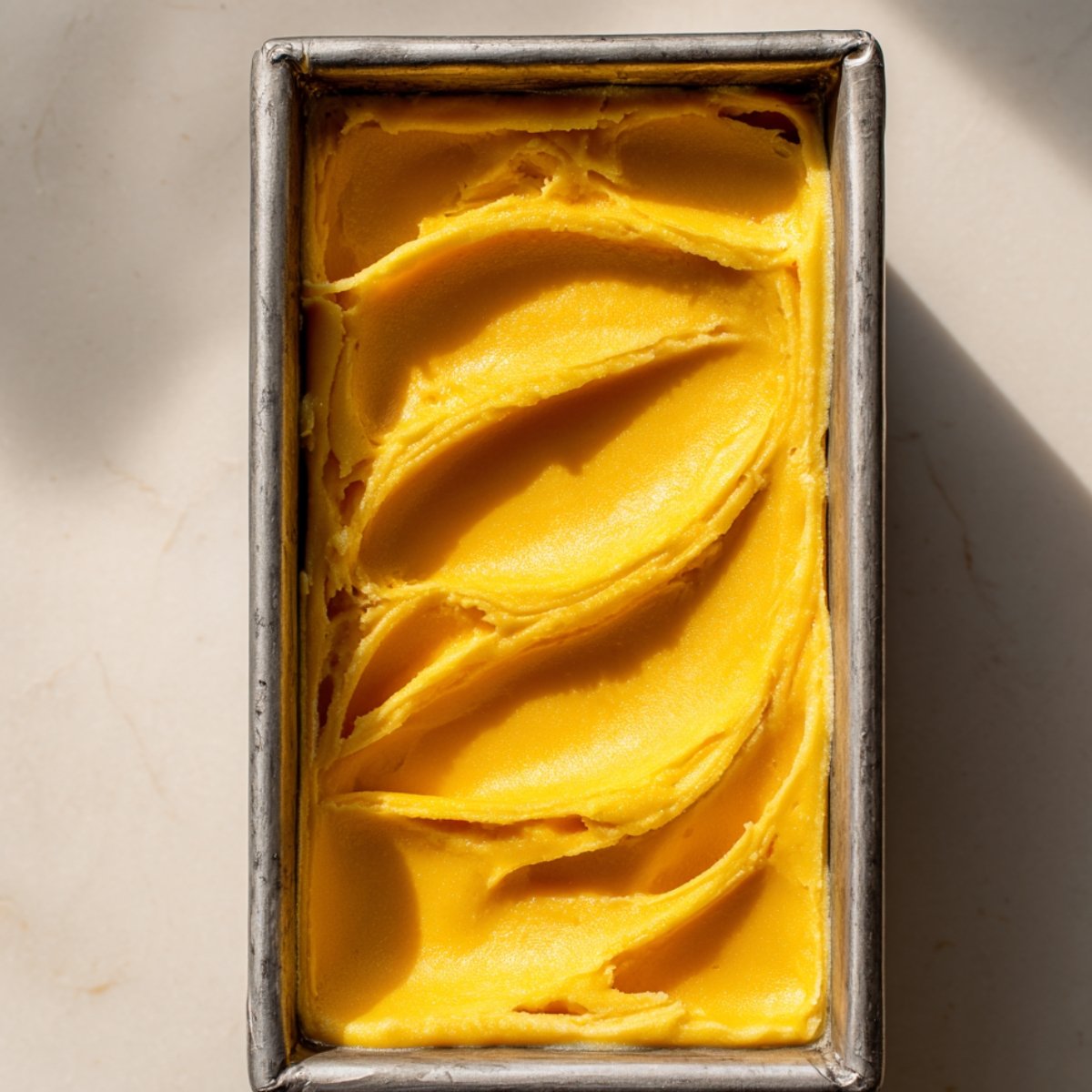 Freshly made mango sorbet in a metal loaf pan, smoothed with soft swirls across the surface. The vibrant yellow-orange sorbet sits on a light marble surface under natural sunlight.