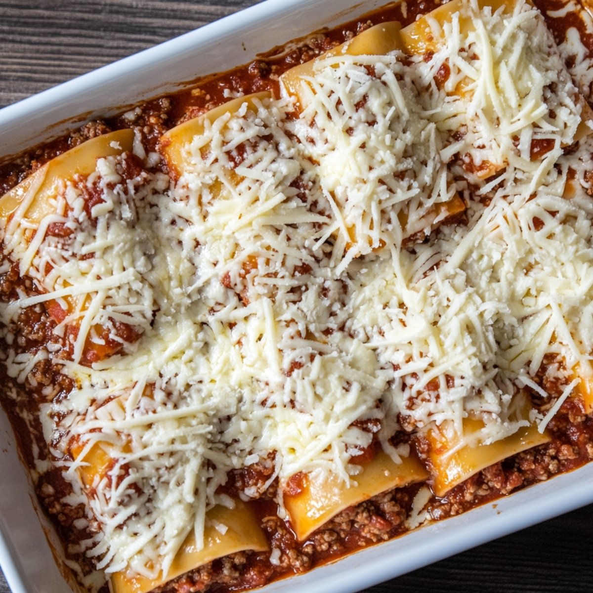 Lasagna roll ups in a white baking dish, topped with shredded mozzarella and nestled in meat marinara sauce, ready to bake.
