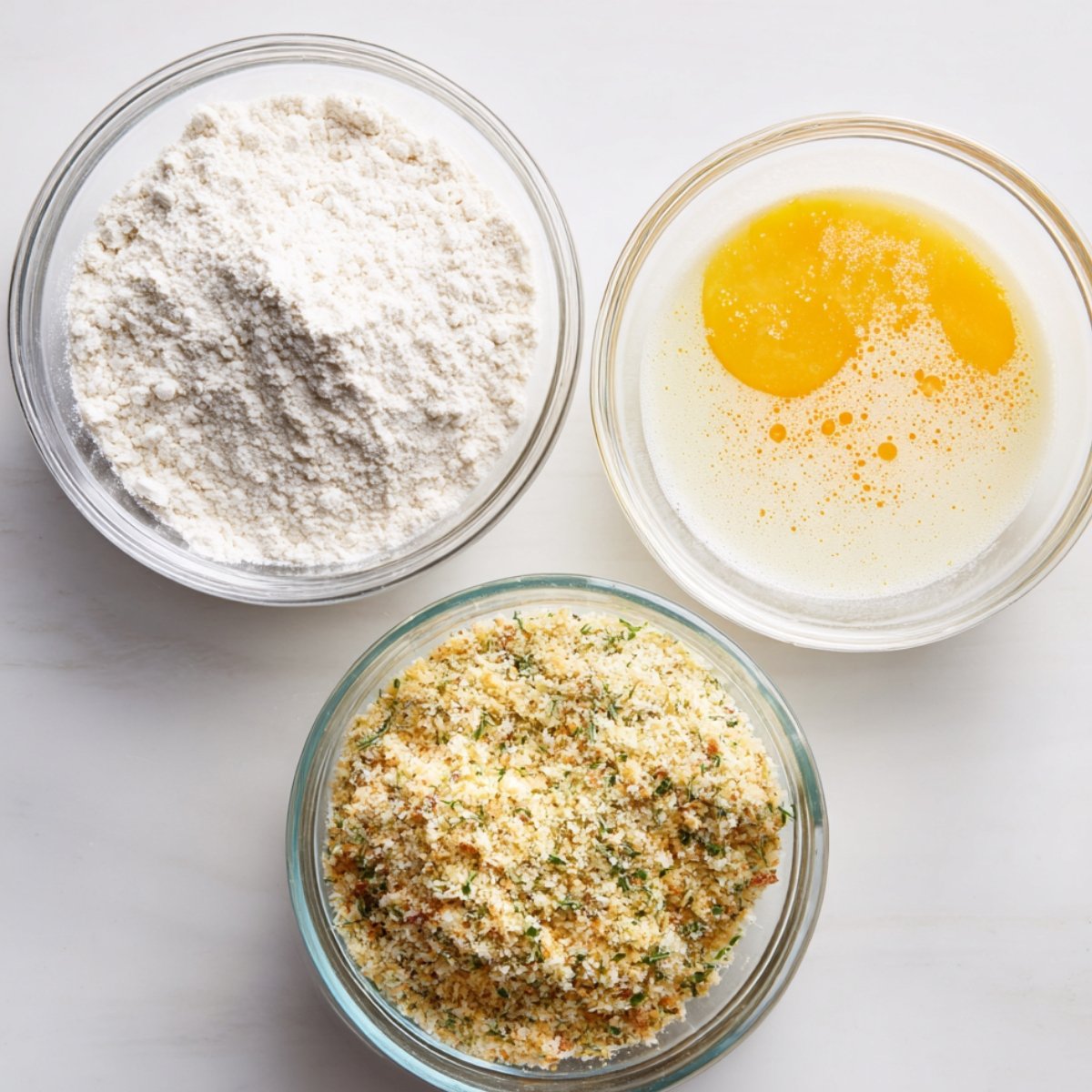 Flour, beaten eggs, and herbed breadcrumbs in glass bowls on a white surface, ready for coating mozzarella sticks.