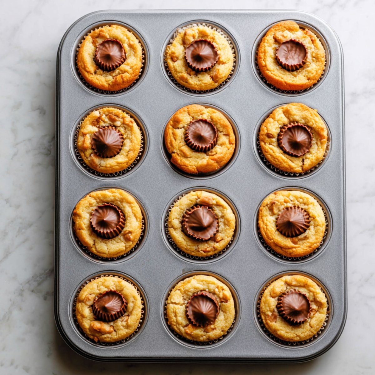 Freshly baked peanut butter cup cookies in a muffin tin, each golden cookie topped with a slightly melted mini peanut butter cup pressed into the center, still warm from the oven.