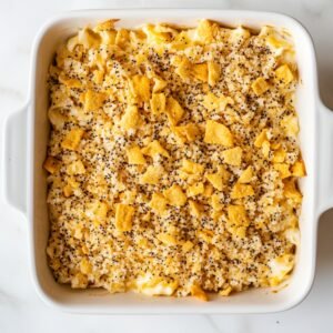 Top-down view of an unbaked poppy seed chicken casserole in a white square baking dish. The creamy chicken mixture is topped evenly with a golden, crushed cracker and poppy seed topping, ready to go into the oven. The dish rests on a clean white marble surface.
