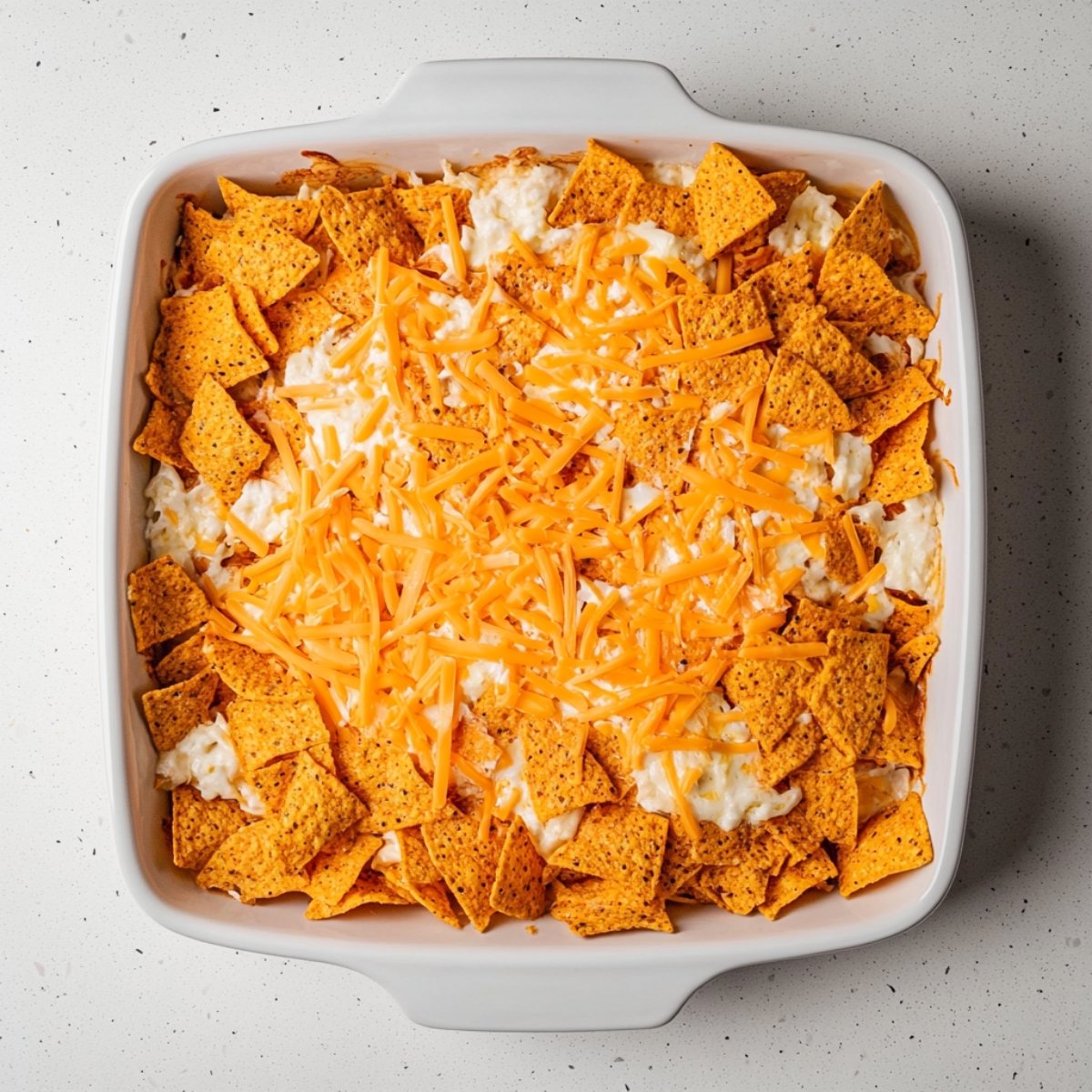 Unbaked Dorito casserole in a white square baking dish, topped with crushed nacho cheese chips and shredded cheddar over a creamy white cheese layer, ready for the oven.
