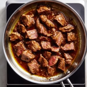 Top-down view of homemade Chinese beef searing in a stainless steel skillet on a black induction cooktop, with tender strips in glossy brown garlic sauce, some pieces still slightly pink, all glistening under natural light.