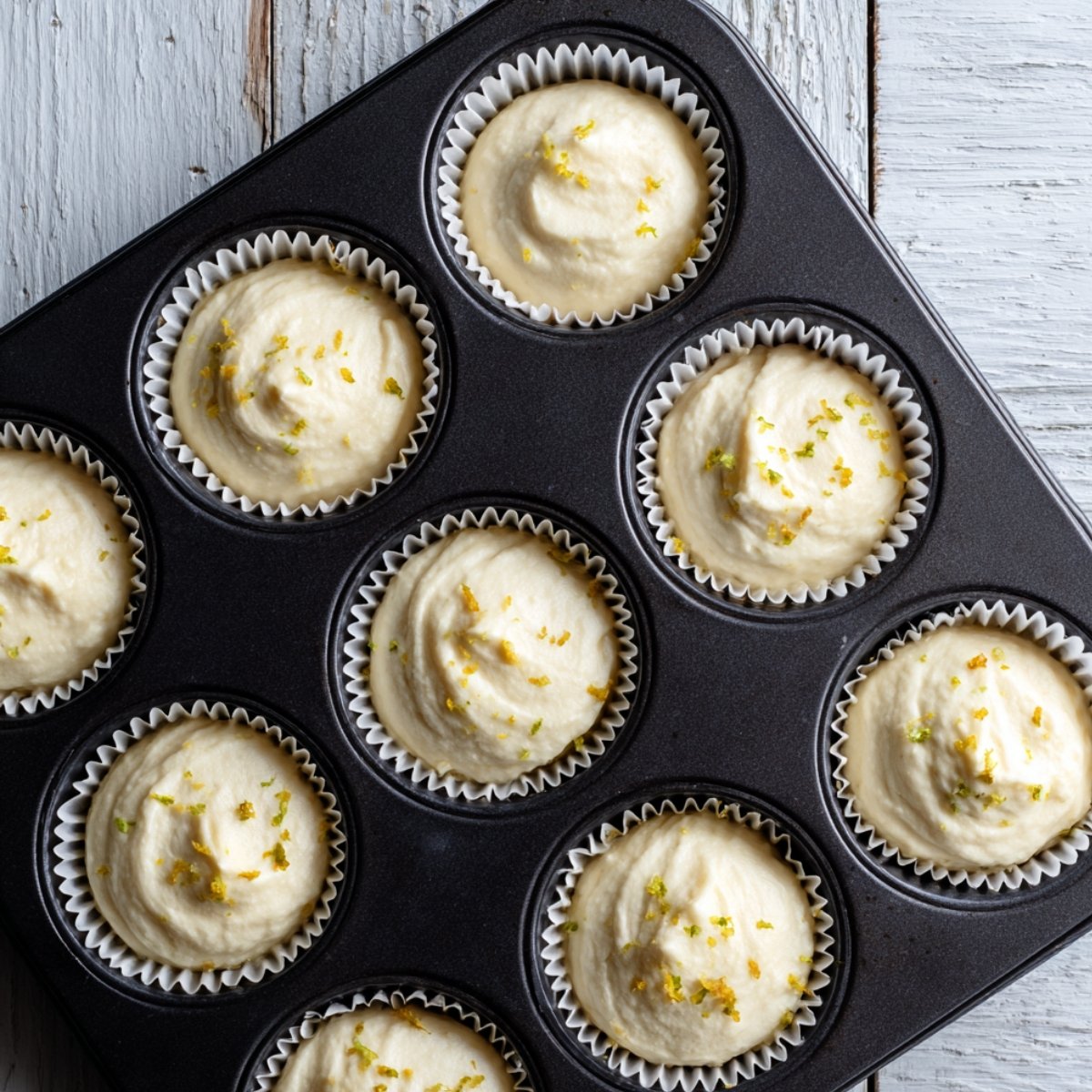 Unbaked raspberry lemon cupcake batter in white paper liners inside a dark muffin tin, topped with fresh lemon zest, ready for the oven. A rustic, homemade kitchen scene.