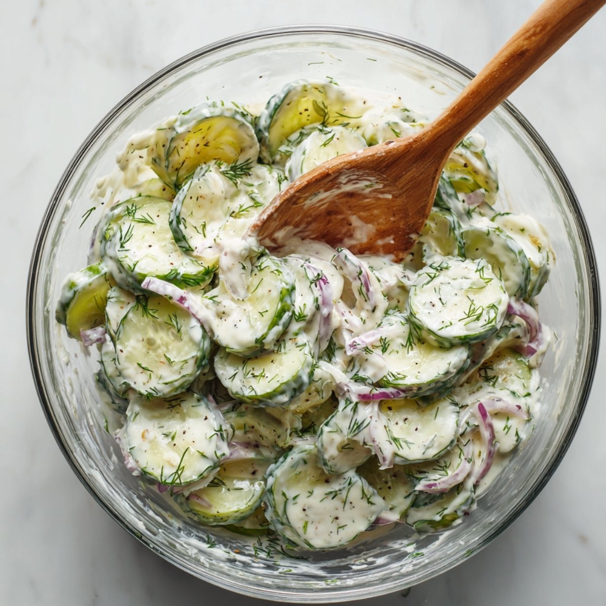 Homemade creamy cucumber salad with fresh dill and red onion in a glass bowl, stirred with a wooden spoon.